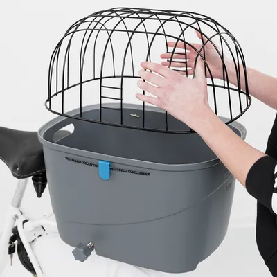 Panier gris en plastique fixé à un vélo, avec une grille de protection noire en métal tenue par une personne au-dessus du panier. Poignée bleue visible sur le devant.