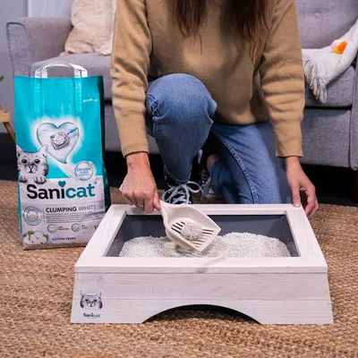 Sanicat Clumping White stelivo pre mačky, osoba odstraňuje hrudky lopatkou z drevenej mačacej toalety. Balenie zobrazuje „Oxygen Power“ a vôňu „Cotton Fresh“.