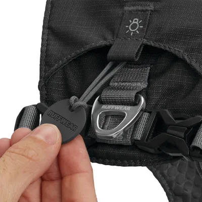 Ruffwear márkájú fekete kutyahám részlete, látható fém csat, műanyag csat és Ruffwear feliratú címke, kéz tartja a címkét.