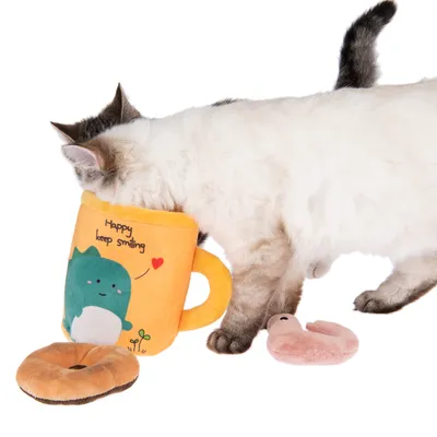 Kočka si hraje se žlutou plyšovou hračkou ve tvaru šálku s nápisem 'Happy keep smiling', vedle plyšový donut a růžová červí hračka.