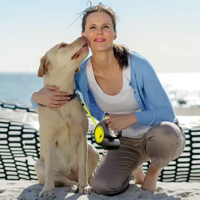 Osoba trzyma psa na żółtej, automatycznej smyczy z czarnym uchwytem podczas spaceru nad morzem. Smycz z widocznym okrągłym elementem i symbolem psa.