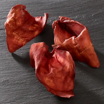 Three dried, reddish-brown animal ear chews displayed on a dark surface, shown whole and unwrapped for product identification.