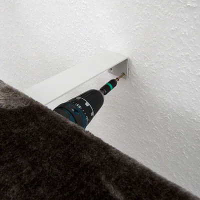 Montageanleitung: Bohrer befestigt eine Schraube an einer Wandhalterung für ein Möbelstück mit grauem Stoffbezug.