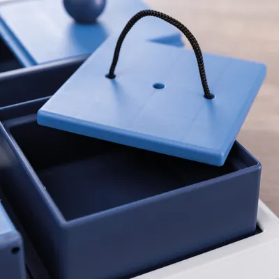 Vassoio in plastica blu con coperchio rimovibile e laccetto nero, parte di un gioco interattivo.