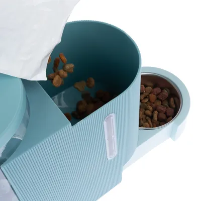 Dry pet food being poured from a bag into a blue automatic feeder with a built-in metal bowl. Shows how to fill the feeder’s storage compartment.