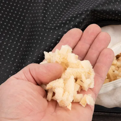 Hand hält kleine Schaumstoffstücke, sichtbar vor einem schwarzen gepunkteten Stoff und einer geöffneten Tasche mit weiteren Schaumstoffstücken.