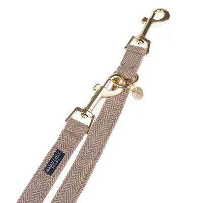 Beige dog lead with herringbone pattern, gold-coloured metal fastenings and pendant. Brand label 'Nomad Tales' visible.