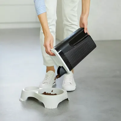 Person pouring dry pet food from a storage container into a bowl with a separate compartment, demonstrating use of the container for dispensing food.