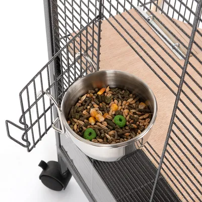 Gamelle en métal accrochée à une cage, remplie de graines, maïs et croquettes vertes pour oiseaux ou petits animaux. Support métallique visible sur la porte de la cage.
