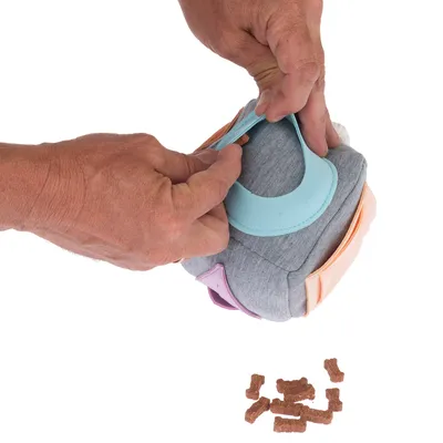 Main ouvrant un jouet interactif en tissu gris avec rabats colorés, friandises en forme d’os visibles à côté sur fond blanc. Aucun texte ou marque visible.