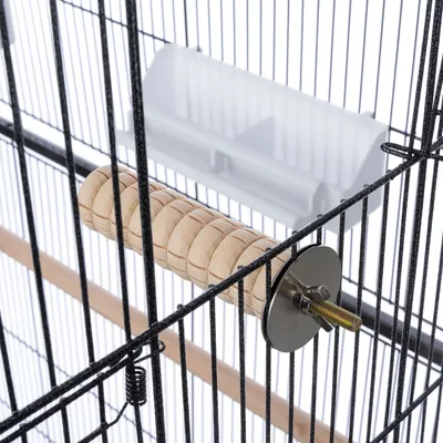 Percha de madera con soporte metálico en una jaula para pájaros, junto a un comedero blanco.