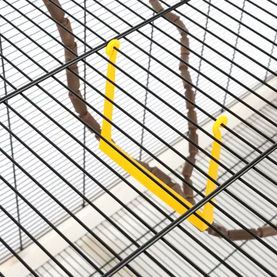 Support jaune en plastique accroché aux barreaux d'une cage à oiseaux, branche en bois visible à l'intérieur de la cage.