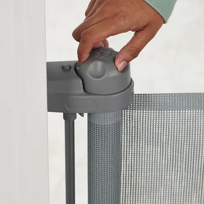 Barrière de sécurité en plastique et maille grise, main actionnant le mécanisme d’ouverture sur le dessus du montant fixé à un mur blanc.