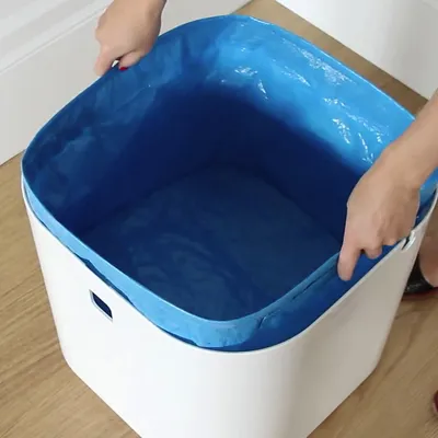 Witte kunststof bak met blauwe binnenzak, vastgehouden aan beide zijden door twee handen. Vierkante vorm, handgrepen zichtbaar aan de zijkanten.