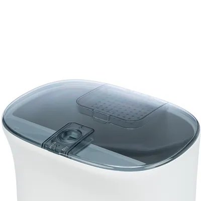 Bílý krmný automat s průhledným víkem a integrovaným dávkovačem na suché krmivo. Viditelný otvor pro dávkování a větrací otvory ve víku.