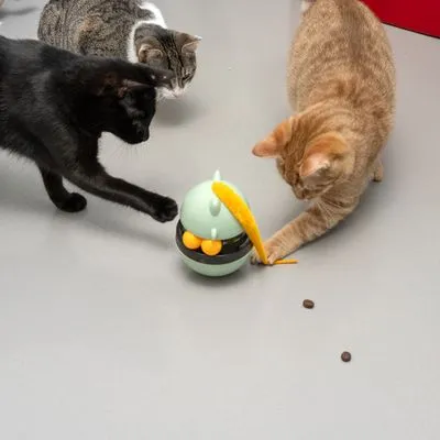 Drie katten spelen met interactief kattenspeelgoed met gele veer en voerbeloning; brokjes liggen op de vloer naast het speelgoed.