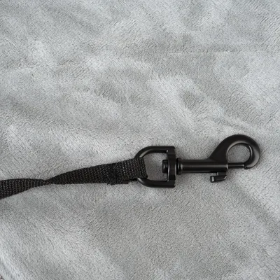 Mousqueton en métal noir fixé à une sangle en tissu noir, posé sur un fond gris. Aucun texte ou marque visible.