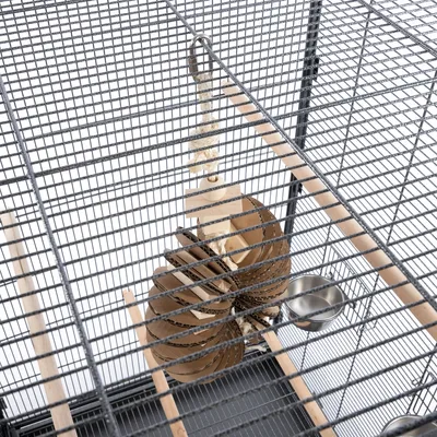 Cage en métal avec perchoirs en bois, jouet suspendu en corde, carton et bois, et deux gamelles métalliques fixées aux barreaux.
