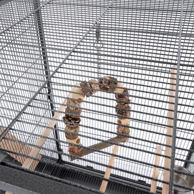 Cage en métal avec balançoire suspendue en bois et éléments naturels, perchoirs en bois et gamelle métallique visibles à l’intérieur.