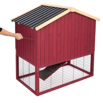 Rotes Holzgehege mit schwarzem Dach und Gitterwänden. Hand öffnet die Tür des oberen Bereichs, Zugang über Rampe sichtbar.
