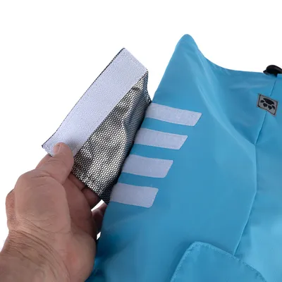 Vue rapprochée d’une main ouvrant une fermeture à scratch sur un manteau bleu pour animal, détails réfléchissants visibles et écusson avec tête de chien.
