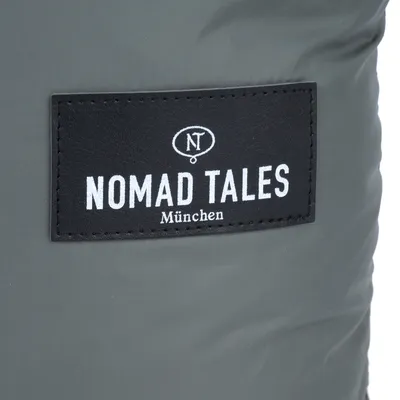 Sort mærke med teksten 'NOMAD TALES München' og et rundt symbol med bogstaverne 'NT' på gråt stof.