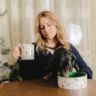 Chien noir mangeant dans une gamelle blanche à motifs de renards, à côté d'une personne tenant une tasse assortie, sur une table en bois près d'un sapin décoratif.