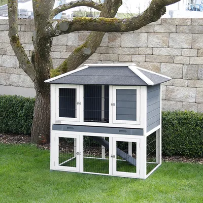Jaula para pequeños animales de dos pisos en gris y blanco con puertas de rejilla, techo inclinado y rampa, colocada en el jardín.