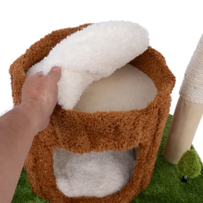 Una mano levanta un cojín blanco extraíble de una cama cueva marrón y mullida para gatos de dos niveles. Rascador de madera y sisal visible al lado.