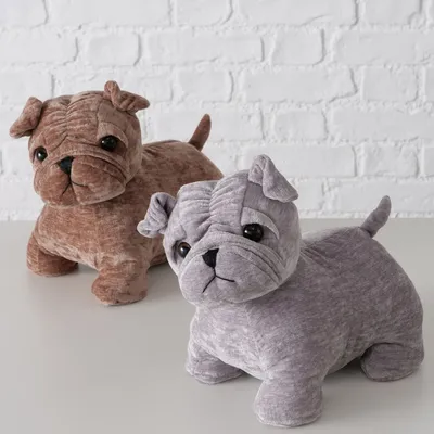 Due cani di peluche marrone e grigio con tratti del viso dettagliati, posizione eretta e coda corta su superficie bianca davanti a un muro di mattoni.