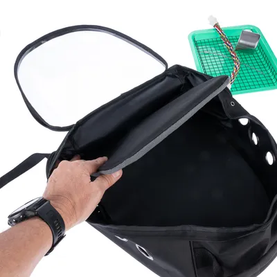 Sac de transport noir ouvert avec poignée, trous d’aération et rabat soulevé. À côté, plateau vert avec grille, corde colorée et gamelle métallique.