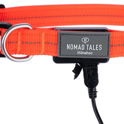 Collier orange en tissu avec boucle métallique, étiquette visible : NOMAD TALES München, câble USB branché sur le collier.