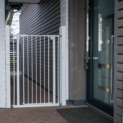 Barrière de sécurité en métal blanc installée entre deux murs à l’extérieur, avec barreaux verticaux et système de fixation visible aux extrémités.