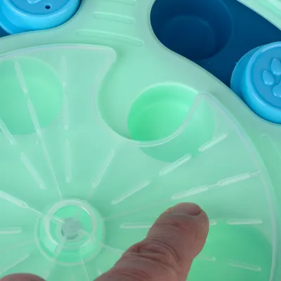 Interactive toy with green and blue surface, finger points to movable element. Promotes dexterity through hidden compartments.