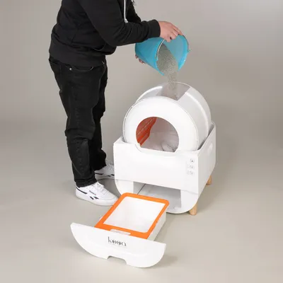Person pouring cat litter from a blue bucket into a white automatic cat toilet with pull-out waste container. kooa brand visible on container.