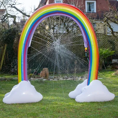 Oppustelig regnbue-vandsprinkler med hvide skyer, sprøjter vandstråler i en have.