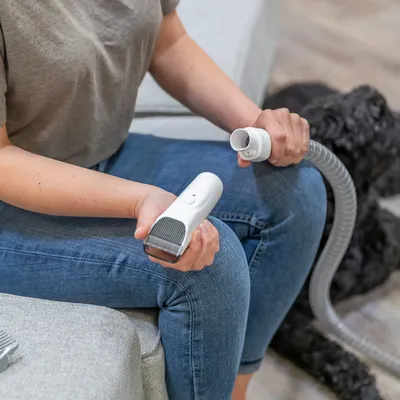 Persona sostiene un cepillo blanco para el cuidado de mascotas con conexión para manguera, adecuado para eliminar pelo de animales. Perro visible al fondo.