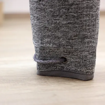 Détail d’un poteau recouvert de tissu gris avec corde tressée insérée dans un œillet métallique à la base, posé sur un sol en bois clair.