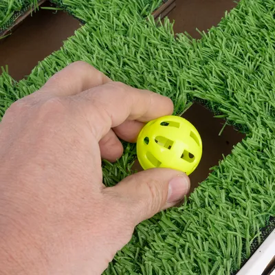 Hand places a yellow plastic ball on a green surface with artificial grass.