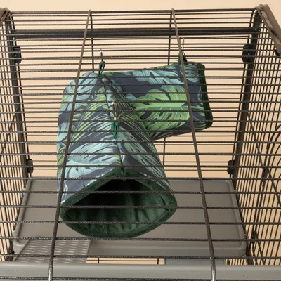 Túnel colgante de tela verde con estampado de hojas, fijado a jaula metálica con ganchos. Adecuado como zona de juego o descanso para pequeños animales.