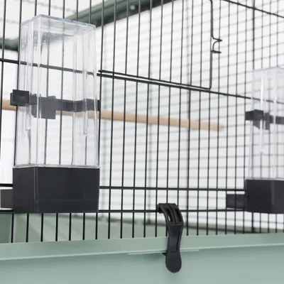 Vogelkooi met transparante voederdispenser en zwarte clip-sluiting, bevestigd aan metalen tralies. Binnenin is een houten zitstok zichtbaar.