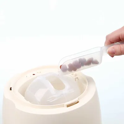 Plastová odměrka s granulemi se sype do otvoru automatického dávkovače krmiva. Viditelná ruka drží odměrku nad zařízením.