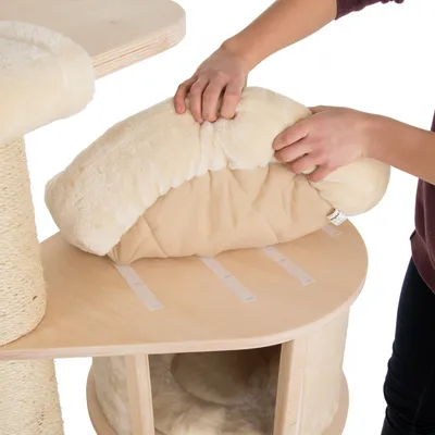 Person befestigt ein weiches, beiges Kissen mit Klettstreifen auf einer Holzplattform eines Kratzbaums.