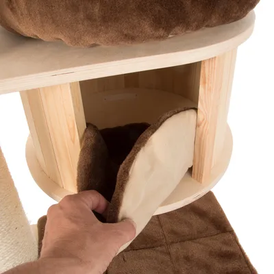 Hand opent ronde kattenhuis van hout met zachte bruine stoffen ingang en binnenkant. Bovenop ligt een bijpassend bruin kussen. Materialen: hout, stof.