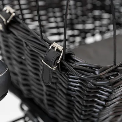 Panier en osier noir avec sangle en cuir noir et boucle métallique, fixé à une structure métallique. Partie d’un siège visible à gauche.