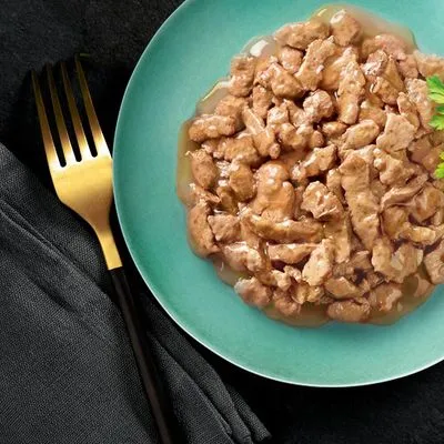 Bord met stukjes natvoer in saus, geserveerd op een groen bord naast een gouden vork en zwarte servet. Merknaam of productinformatie niet zichtbaar.