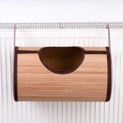 Niche suspendue en bambou avec ouverture sur le dessus, accrochée à un radiateur blanc. Structure cylindrique, bords renforcés en tissu foncé.