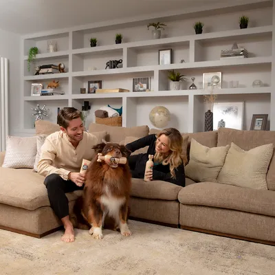 Cane tiene un gioco da masticare a forma di osso, mentre due persone sedute su un divano tengono prodotti per la cura del cane.