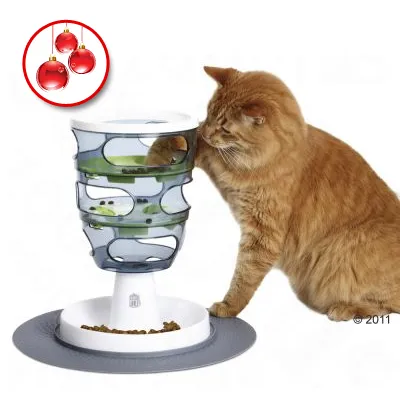 Kissa käyttää muovista aktivointiruokinta-automaattia, jossa on useita tasoja ja reikiä. Alustalla kissanruokaa. Vasemmassa yläkulmassa kolme punaista joulupalloa.