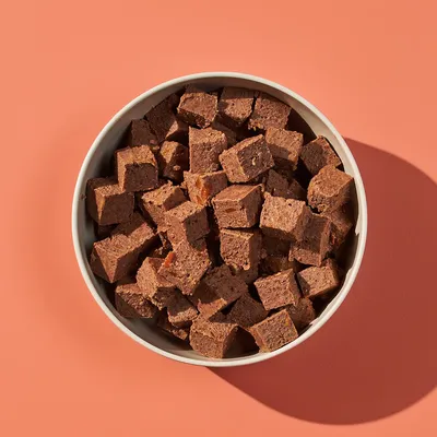 Bowl filled with brown, cube-shaped pet food treats or snacks, shown from above on a plain background.
